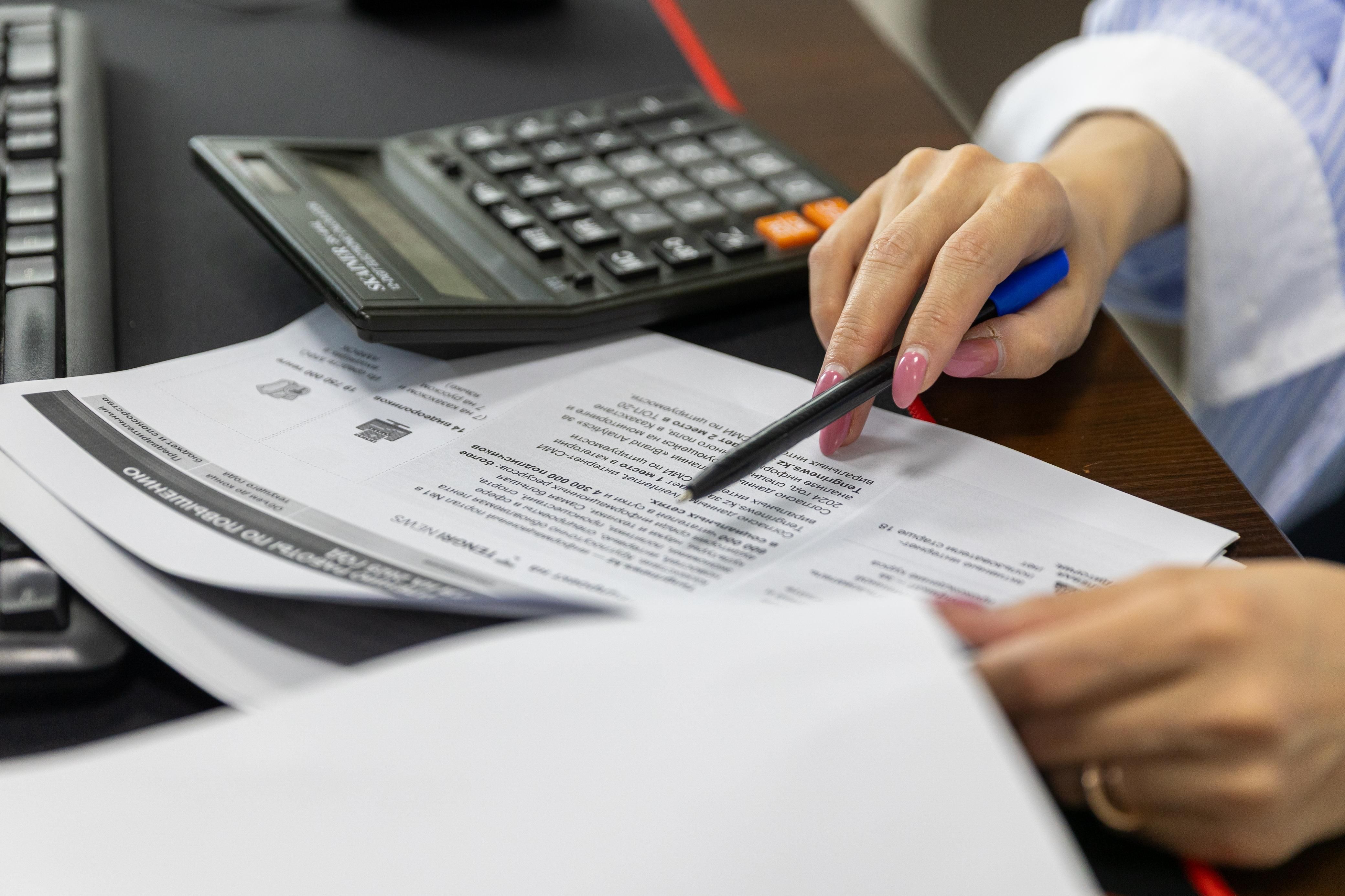 Person with pink nails pointing at document text with pen near calculator on desk.