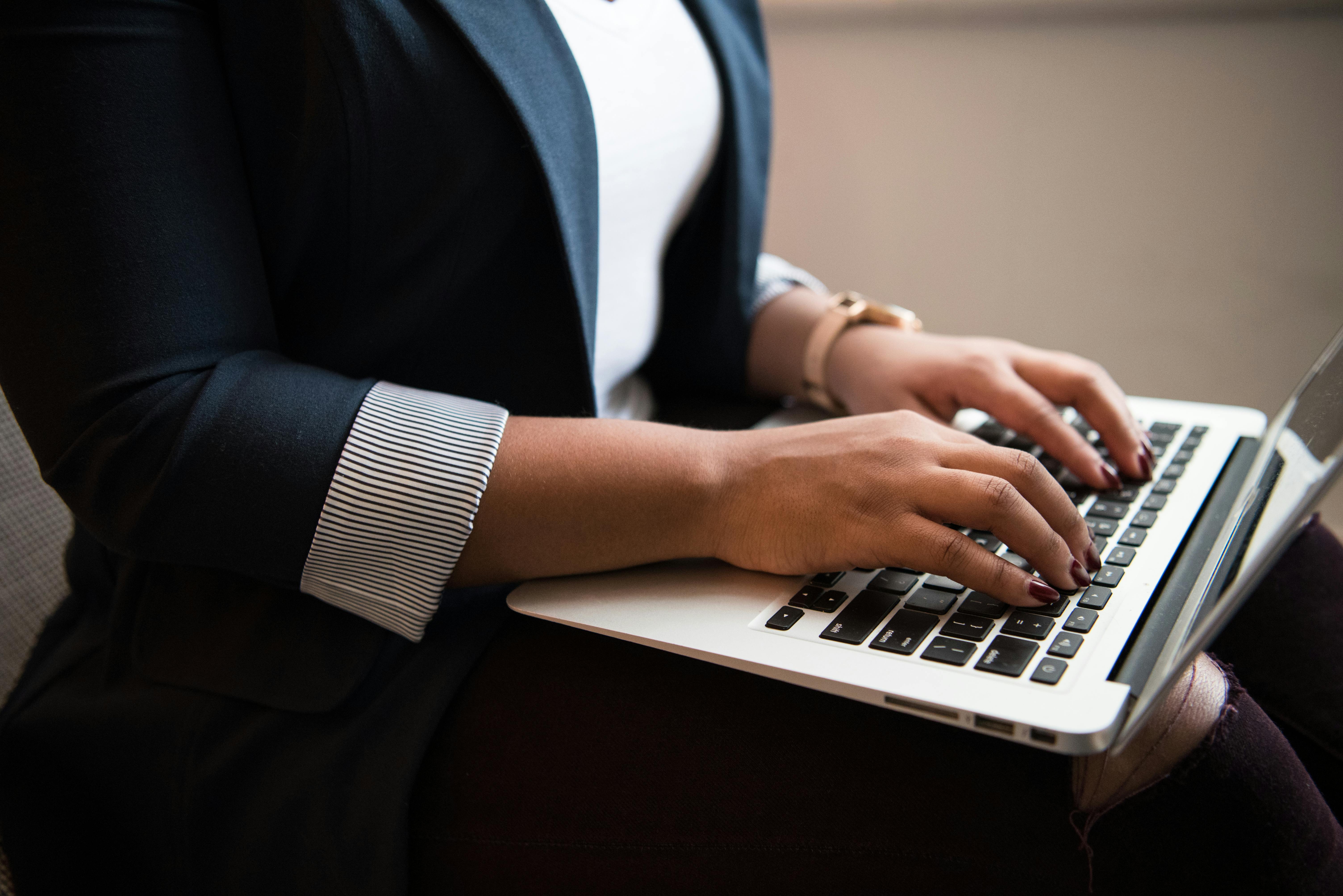 Person in business attire typing on a laptop with dark nails and a watch on their wrist.
