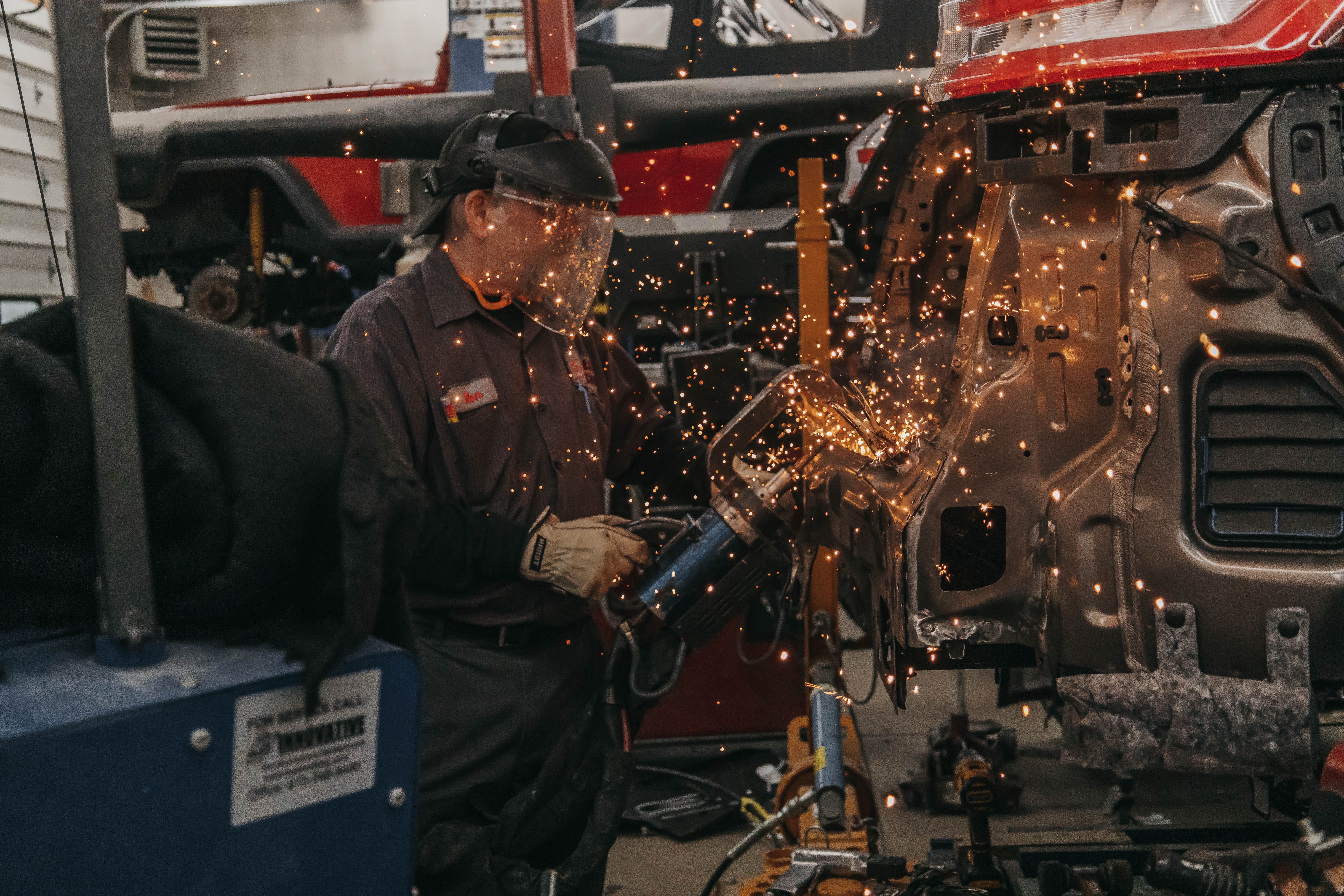 Worker wearing face shield grinding metal on a vehicle, producing bright sparks in a workshop.