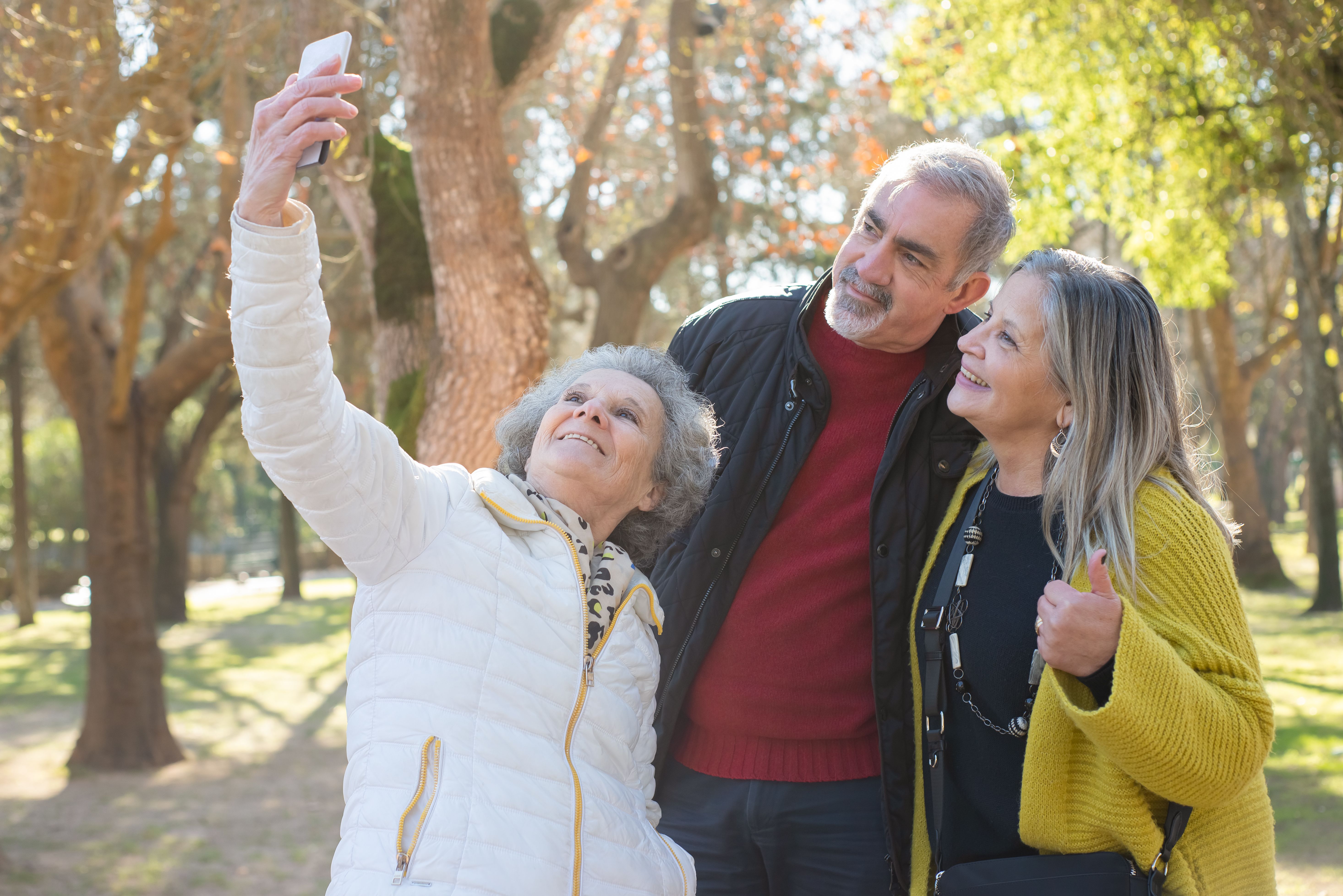 Three smiling adults outdoors taking a selfie in a sunlit park with trees in the background.