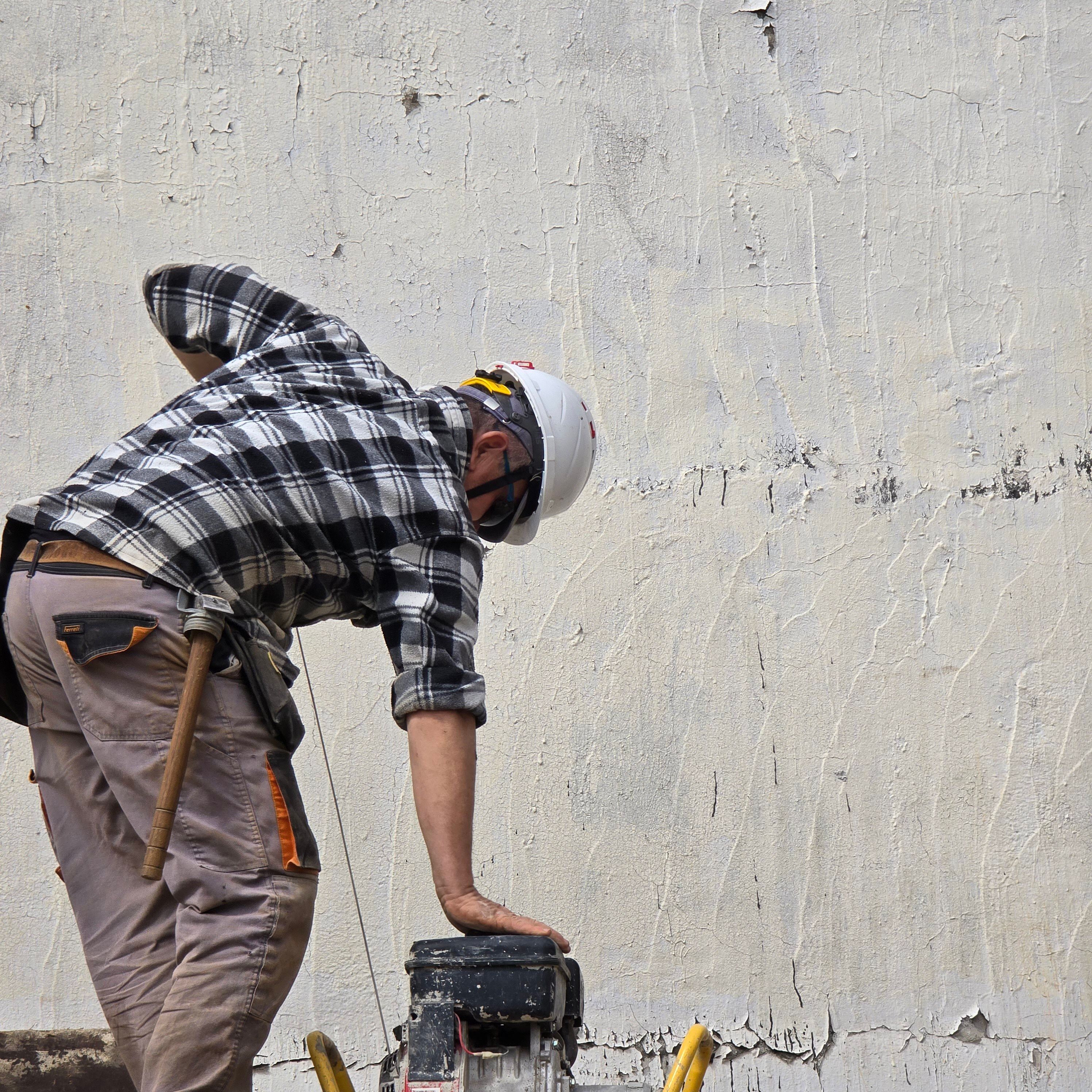 Worker in plaid shirt and helmet operating a machine by a cracked, rough wall surface.