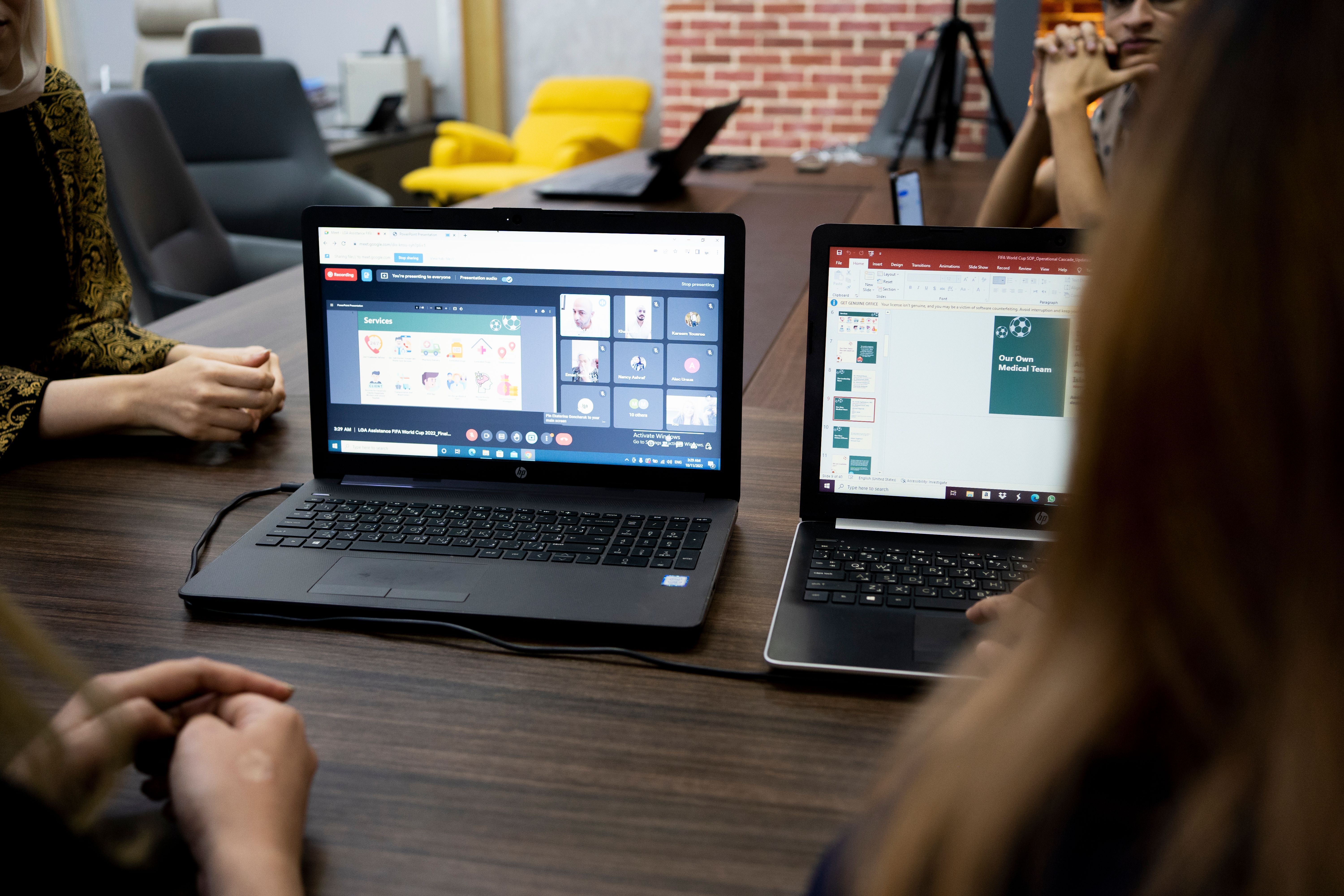 Two laptops on table showing online meeting and presentation with people around the table.