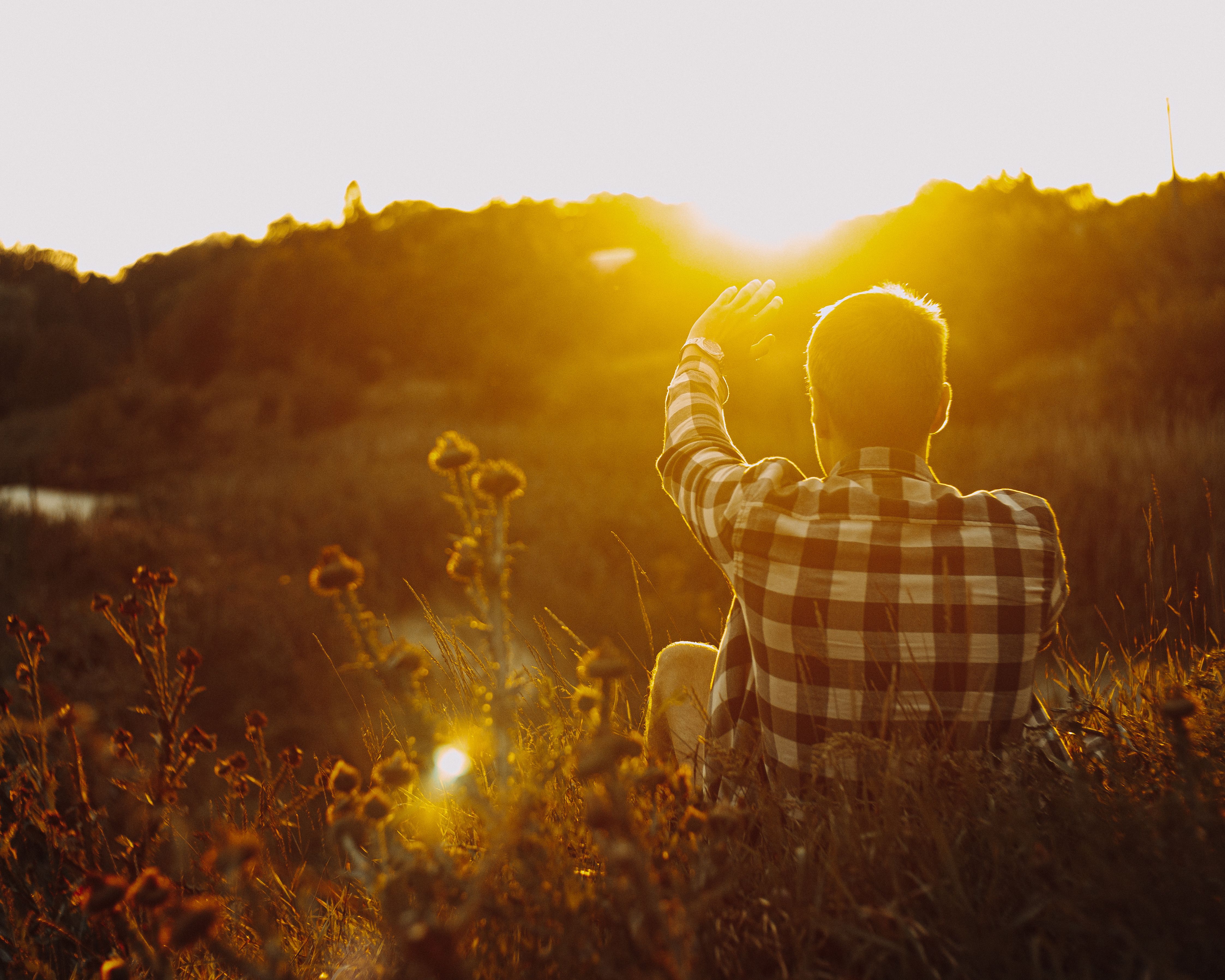Person in plaid shirt sitting in a field waving toward the bright setting sun.