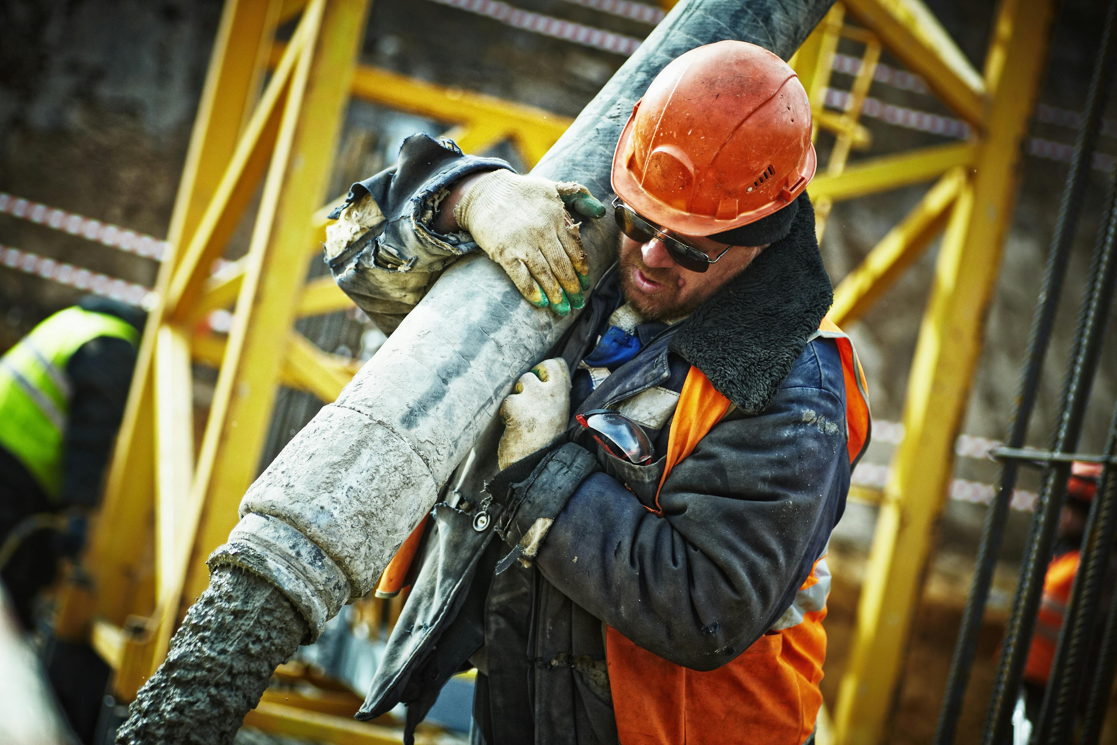 Construction worker in helmet and gloves pouring concrete through large hose at site.